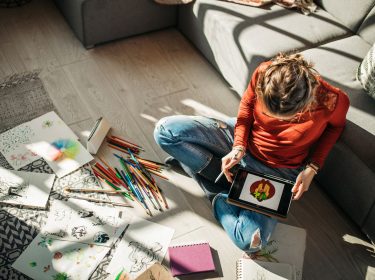 Eine Frau sitzt auf dem Wohnzimmerboden und zeichnet via Tablet.