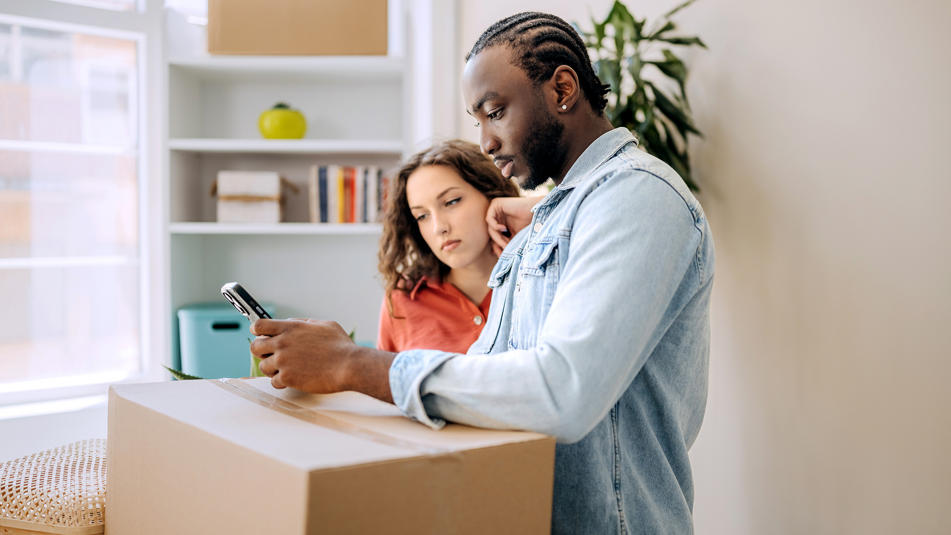 Auf dem Bild in dem Artikel zum Umzugsservice von Vodafone stehen ein junger Mann und eine Frau mit lockigem Haar vor einem großen Umzugskarton. Beide schauen konzentriert auf das Smartphone des Mannes.