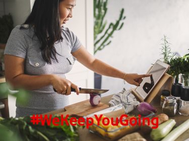 Eine Frau schneidet Gemüse in der Küche und nutzt ihr Tablet, um ein Gericht per Anleitung zuzubereiten.