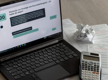 Ein Laptop, auf dessen Bildschirm der Chatbot UDO zur Beantragung von Kurzarbeitergeld zu sehen ist, daneben Unterlagen und ein Taschenrechner.