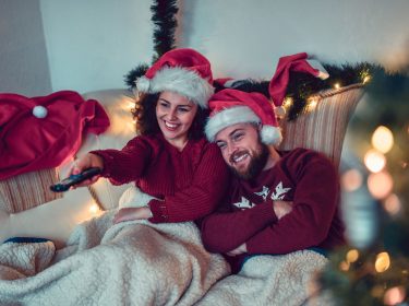 Ein junges Paar mit Weihnachtspulli und -mütze schaut zusammen Fernsehen.