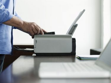 Laptop im Vordergrund, im Hintergrund arbeitet ein Mann am Drucker