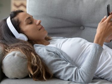 Frau mit Bluetooth-Kopfhörern liegt auf dem Sofa und nimmt Einstellungen am Handy vor