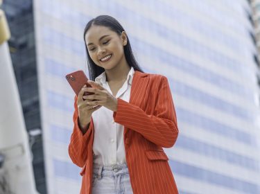 Frau mit rotem Blazer schaut lächelnd auf ihr iPhone.