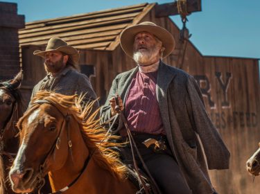 Im Artikel zu den besten Western-Serien siehst du ein Bild aus der Serie Godless ist Jeff Daniels als Frank Griffin. Er reitet auf einem braunen Pferd durch eine staubige Westernstadt, begleitet von einem weiteren Reiter