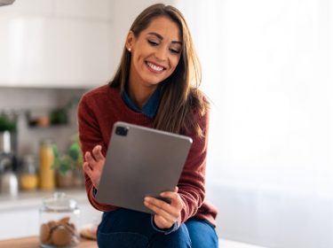 Lächelnde Frau mit iPad Pro sitzt auf dem Küchentisch.