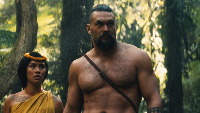 Auf dem Bild aus der Serie Chief of War ist Jason Momoa in einer zentralen Szene im Dschungel zu sehen. Er steht oberkörperfrei neben einer weiteren Figur, beide wirken angespannt und fokussiert. Die Szene ist atmosphärisch dicht, mit dramatischer Beleuchtung im tropischen Wald. Ideal für Fans von Jason Momoas beste Filme und Serien.