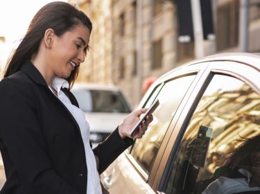 Frau öffnet Tür eines Autos, während sie ihr Smartphone in der Hand hält.
