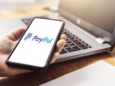 Eine Hand hält ein Smartphone-Display mit dem PayPal-Logo vor einen Laptop.