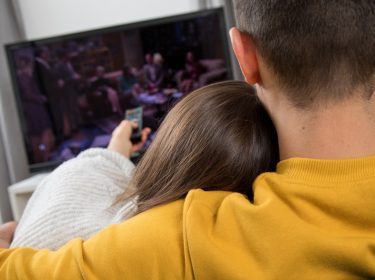 Junge Frau und junger Mann sitzen Arm in Arm auf dem Sofa und gucken Fernsehen