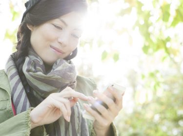 Frau mit Sonne und Baum im Rücken guckt lächelnd auf ihr iPhone