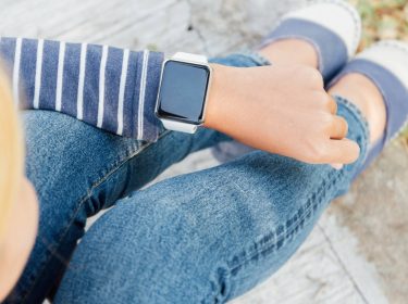 Blonde Frau sitzt auf Brüstung und schaut auf Apple Watch am Handgelenk
