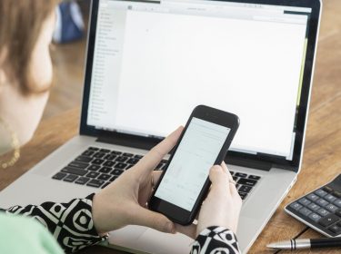 Frau sitzt vor Laptop und hält Smartphone in beiden Händen