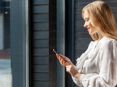 Junge Frau vor Fenster blickt lächelnd auf ihr Smartphone