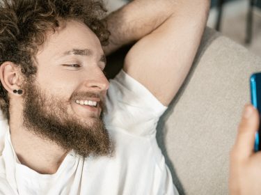Junger Mann mit Bart und verwuschelten Haaren liegt auf der Couch und guckt lächelnd auf sein Smartphone