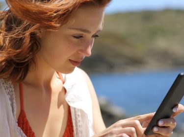 Rothaarige Frau am Meer guckt auf ihr Smartphone