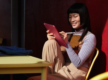 Schwarzhaarige mit pinkem Apple iPad 10 und Apple Pencil arbeitet entspannt an eine Hauswand gelehnt