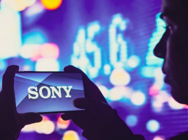 Mann vor bunten Leuchttafeln hält Handy mit beiden Händen, auf dem Display das Sony-Logo
