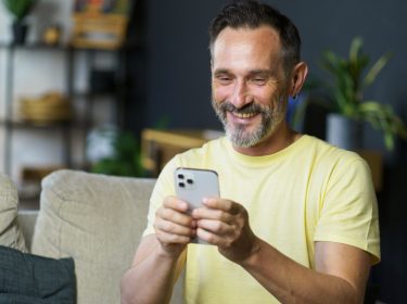 Mann mittleren Alters sitzt lächelnd auf der couch und guckt auf sein iPhone