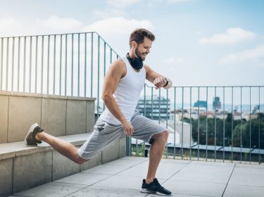 Junger Mann auf Dach trainiert an Stufen aus Stein und schaut lächelnd auf seine Smartwatch
