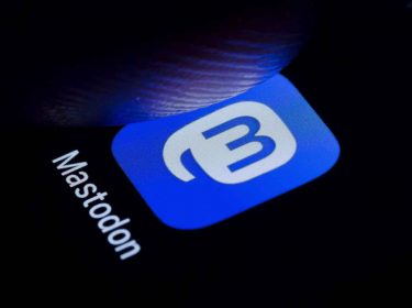 Das App-Logo von Mastodon auf einem Smartphone-Bildschirm, ein Stück über dem Bildschirm befindet sich ein Finger in der Luft