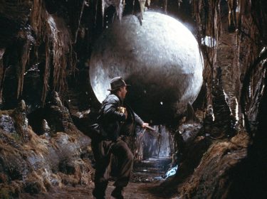 Indiana Jones (Harrison Ford) läuft vor einer Steinkugel weg, die ihn droht zu erdrücken in einem der besten Filme von George Lucas