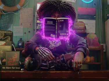 Layla sitzt im Schneidersitz auf einem Tresen und nutzt Telekinese, um ein Buch vor sich schweben zu lassen im Spiel Redfall Kritik