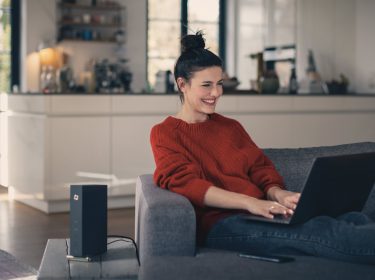 Frau sitzt auf Sofa mit Laptop und Kabelrouter von Vodafone steht auf dem Tisch