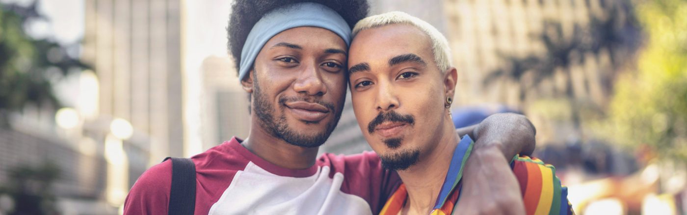 Auf dem Bild zur CSD Vodafone Kampagne umarmen sich zwei Männer und schauen selbstbewusst in die Kamera. Einer trägt ein Hemd in Regenbogenfarben, das Symbol für Pride und Vielfalt. Die Szene spielt in einer belebten Innenstadt mit Hochhäusern und sommerlicher Atmosphäre – ein Zeichen für Zusammenhalt, Sichtbarkeit und Offenheit.