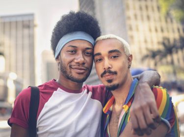 Auf dem Bild zur CSD Vodafone Kampagne umarmen sich zwei Männer und schauen selbstbewusst in die Kamera. Einer trägt ein Hemd in Regenbogenfarben, das Symbol für Pride und Vielfalt. Die Szene spielt in einer belebten Innenstadt mit Hochhäusern und sommerlicher Atmosphäre – ein Zeichen für Zusammenhalt, Sichtbarkeit und Offenheit.