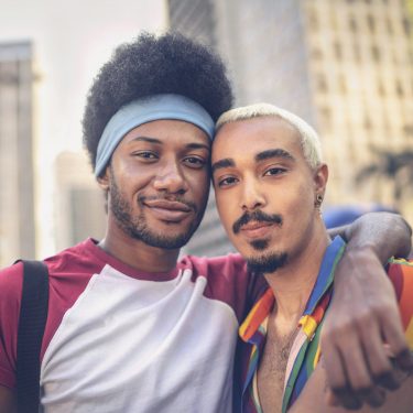 Auf dem Bild zur CSD Vodafone Kampagne umarmen sich zwei Männer und schauen selbstbewusst in die Kamera. Einer trägt ein Hemd in Regenbogenfarben, das Symbol für Pride und Vielfalt. Die Szene spielt in einer belebten Innenstadt mit Hochhäusern und sommerlicher Atmosphäre – ein Zeichen für Zusammenhalt, Sichtbarkeit und Offenheit.