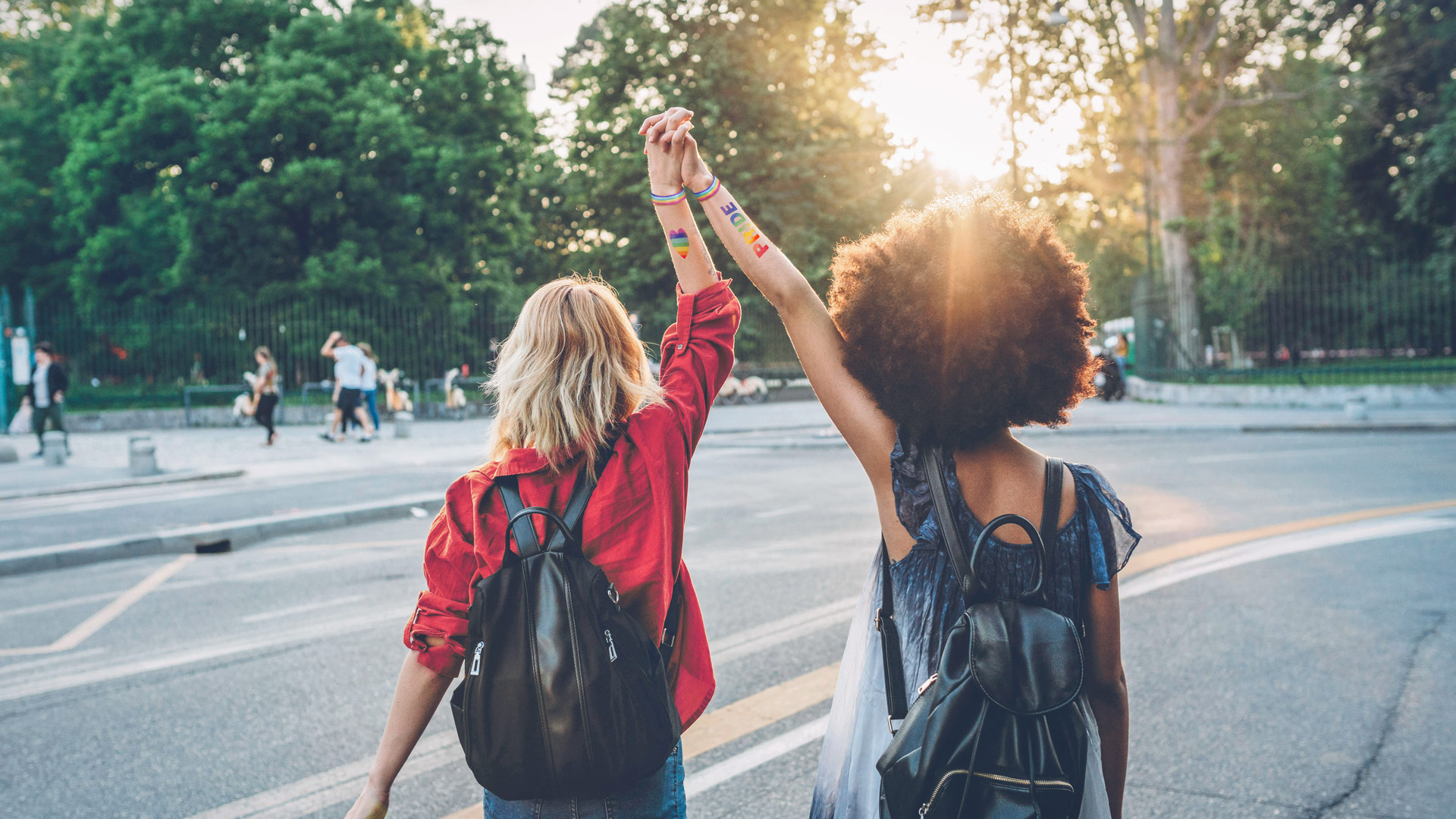 Auf dem Bild zur CSD 2025 mit Vodafone halten zwei junge Frauen mit Rucksäcken und Regenbogenarmbändern ihre Hände hoch. Die Worte „PRIDE“ und ein Herz in Regenbogenfarben sind auf ihren Armen zu sehen. Die Sonne scheint durch die Bäume im Hintergrund, was dem Bild eine optimistische, inklusive und freie Stimmung verleiht.