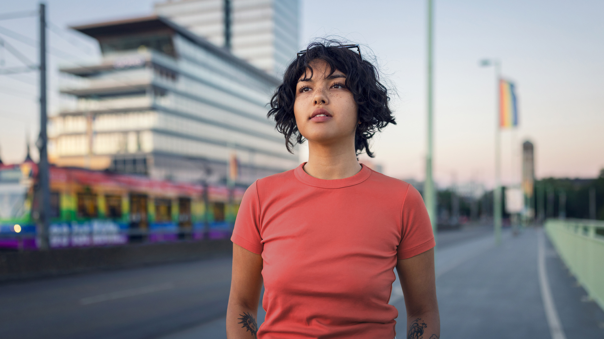 Auf dem Bild zum CSD mit Vodafone steht eine junge Frau auf einer Brücke in Köln. Hinter ihr Fährt eine Bahn in Regenbogenfarben, was das Thema Pride weiter verstärkt.
