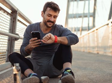 Mann sitzt auf dem Boden, macht Pause vom Sport und schaut lächelnd auf seine Smartwatch und sein Handy