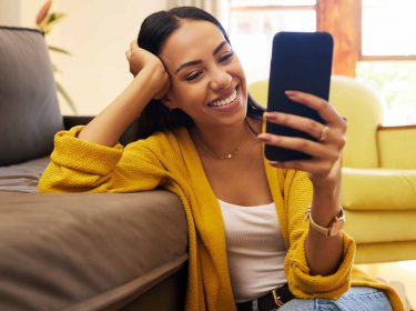 Eine Frau hockt auf dem Boden, hat einen Arm auf der Couch und ein Handy in der Hand
