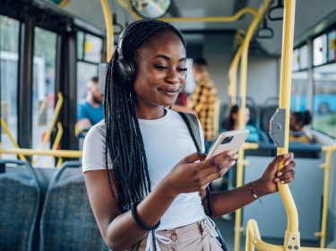 Junge Frau im Bus hört Musik und schaut lächelnd auf ihr Handy