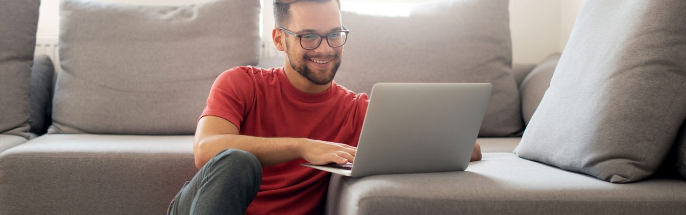 Junger Mann sitzt auf dem Boden vor seinem Sofa und ist lächelnd an seinem Laptop