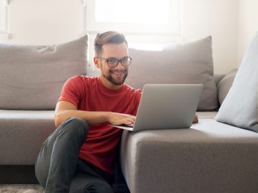 Junger Mann sitzt auf dem Boden vor seinem Sofa und ist lächelnd an seinem Laptop