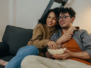 Mann und Frau sitzen lächelnd und kuschelnd auf dem Sofa mit einer Schüssel Popcorn in der Hand