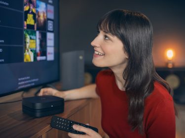 Bei der Auswahl der Vodafone Premium Plus Sender blickt eine junge Frau lächelnd auf den Fernseher, während sie eine Fernbedienung in der Hand hält.