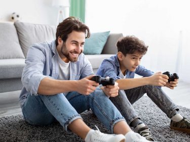 Ein Vater spielt mit seinem Sohn etwas auf einer Console und sitzt dabei auf dem Boden vor einer Couch