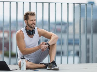 Junger Mann sitzt mit Laptop und Smartwatch auf einer Dachterasse
