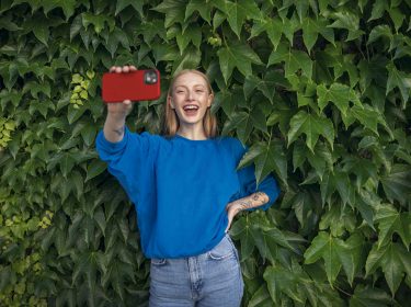 Eine junge Frau steht lächelnd vor einer Efeu-Wand und hält ihr Smartphone in die Kamera