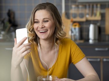 Eine junge Frau sitzt in einer Küche, schaut in ihr Smartphone und lächelt dabei.