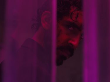 Dev Patel wird rot angestrahlt und steht hinter einem lila Vorhang