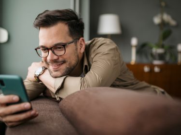 Ein Mann mit Brille liegt lächelnd auf einer Couch und hält ein Smartphone in seinen Händen und nutzt eine der besten kostenlosen Dating-Apps