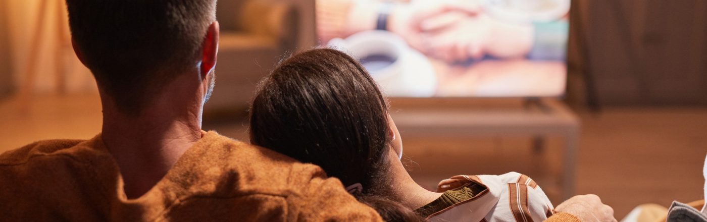 Auf dem Bild in dem Artikel zu den Filmen zum Valentinstag sitzt ein Paar gemütlich auf dem Sofa und schaut gemeinsam einen Film. Der Mann trägt einen braunen Pullover und hat den Arm um die Frau gelegt, die sich an seine Schulter lehnt. Auf dem Fernseher läuft eine verschwommene Szene mit zwei Händen und einer Tasse Kaffee, die eine romantische Stimmung vermittelt.