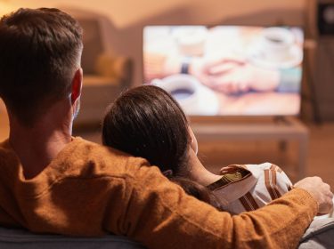 Auf dem Bild in dem Artikel zu den Filmen zum Valentinstag sitzt ein Paar gemütlich auf dem Sofa und schaut gemeinsam einen Film. Der Mann trägt einen braunen Pullover und hat den Arm um die Frau gelegt, die sich an seine Schulter lehnt. Auf dem Fernseher läuft eine verschwommene Szene mit zwei Händen und einer Tasse Kaffee, die eine romantische Stimmung vermittelt.
