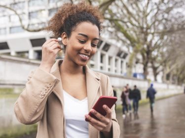 Eine junge Frau mit Smartphone in einer Hand. Mit der anderen Hand steckt sie einen In-Ear-Kopfhörer in ihr Ohr.