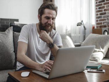 Ein Mann sitzt auf der Couch und nutzt dabei einen Laptop auf dem Tisch vor sich.